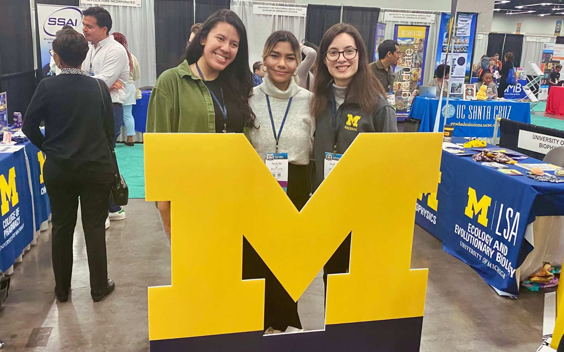 Three women posing in front of a the U-M “block M” sign at a conference.