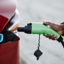 A person in a suit plugs a green electric vehicle charger into a red car, indicating the action of charging the vehicle.
