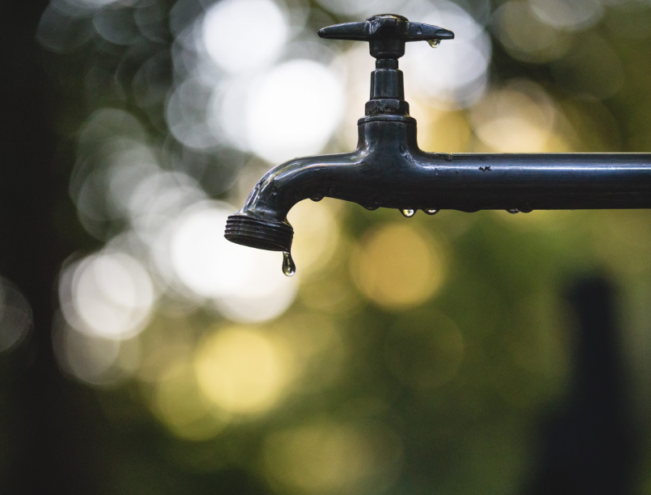 A close-up of a metal faucet with a droplet of water hanging, set against a blurred, nature-inspired background.