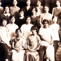 A group of women in early 20th-century attire pose together in three rows against a dark curtain background.