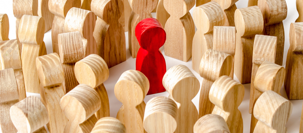 A red wooden figure stands out in the center of numerous natural wood figures, symbolizing a feeling of being judged.