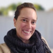 A person with short brown hair wearing a thick dark scarf and beige coat smiles at the camera; a blurred background is visible.