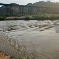 River with rippling water in foreground, bridge and mountainous landscape in background. Sparse vegetation lines the riverbank.