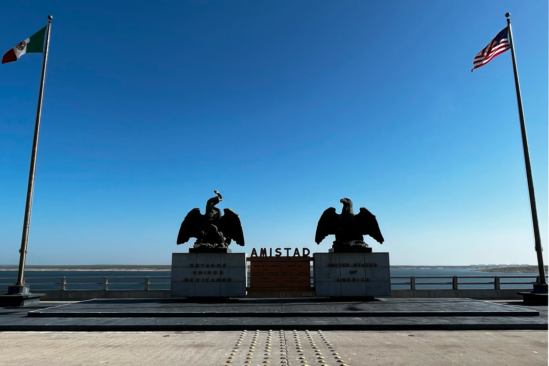 Statues of eagles between Mexican and American flags at Amistad Dam on the US-Mexico border under a clear blue sky.