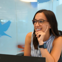 A person sitting at a desk with a laptop smiles, wearing glasses and a smartwatch. Blue bird decals adorn the glass wall behind them.