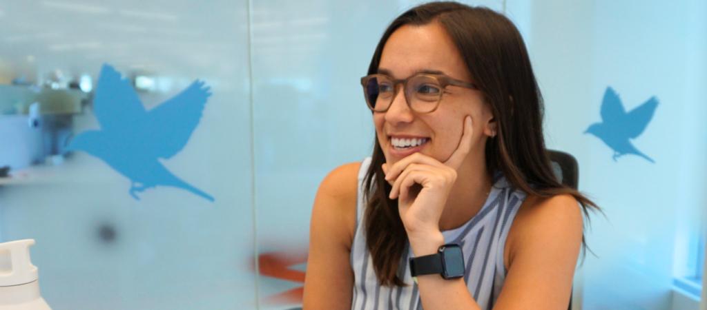 A woman wearing glasses and a sleeveless top is sitting at a table with a laptop and water bottle in an office setting. She is smiling and resting her hand on her chin.