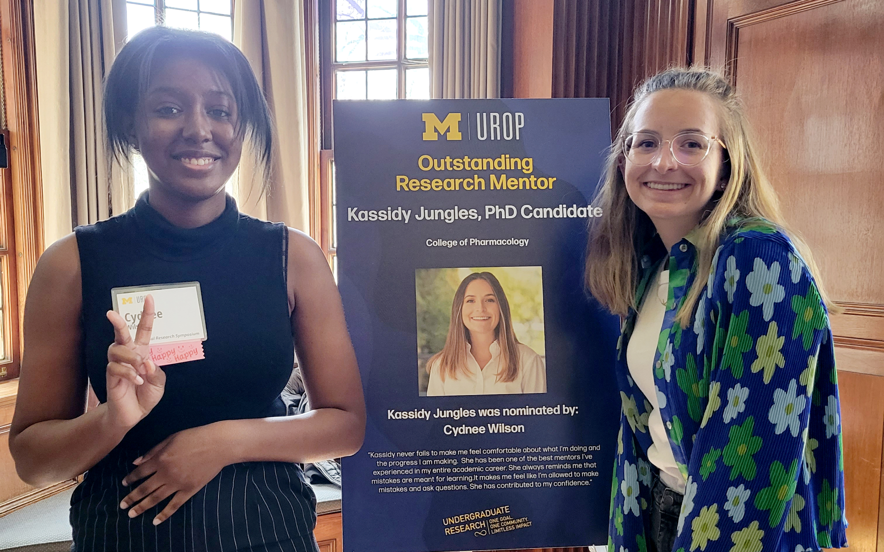 Two people stand next to a sign recognizing Kassidy Jungles, a PhD candidate in Pharmacology, as an outstanding research mentor.