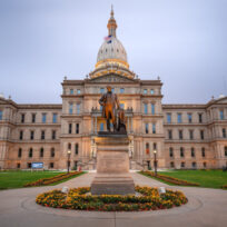 A wide view of the Michigan State Capitol building in Lansing, with a statue of a man on a pedestal in front, surrounded by flowerbeds and a cloudy sky overhead.