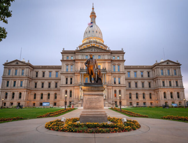 A wide view of the Michigan State Capitol building in Lansing, with a statue of a man on a pedestal in front, surrounded by flowerbeds and a cloudy sky overhead.