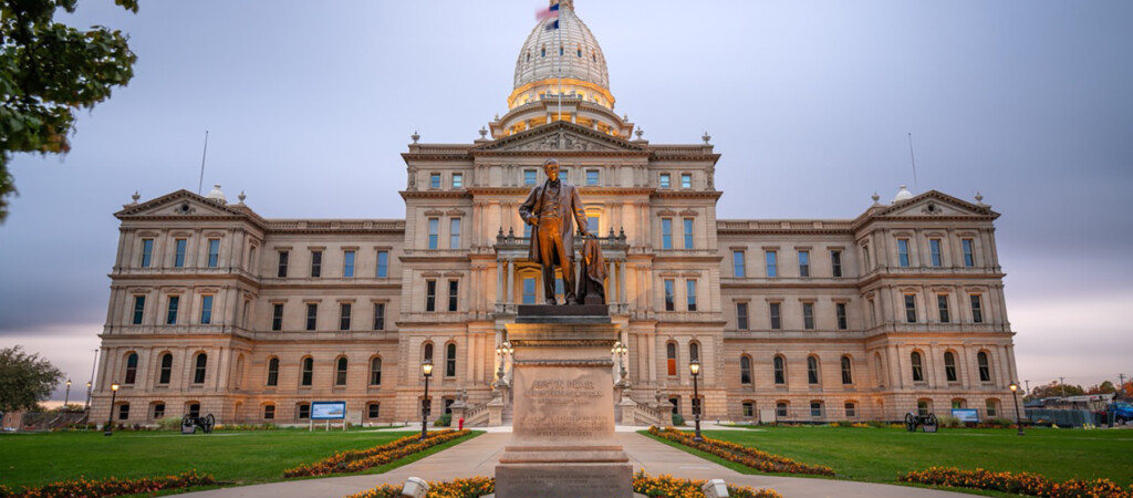 The image shows the exterior of a large neoclassical government building with a dome, surrounded by landscaping and featuring a prominent statue in the foreground.