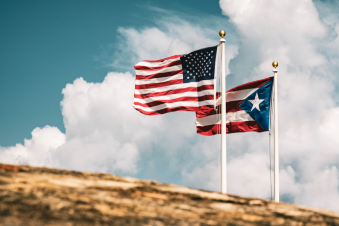 Two flags on poles against a cloudy sky: the United States flag on the left and the Puerto Rico flag on the right.