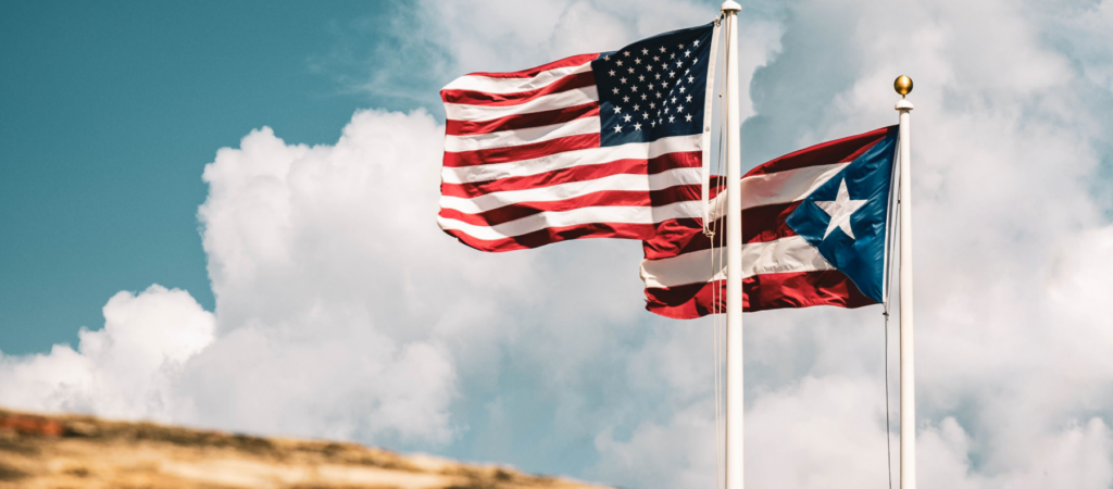 Flags of the United States and Puerto Rico waving on flagpoles against a cloudy blue sky.