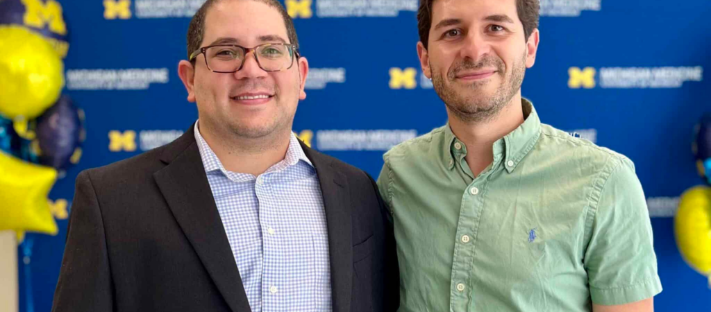 Two men standing in front of a University of Michigan backdrop with balloons. One is wearing a suit, and the other is in a green shirt.