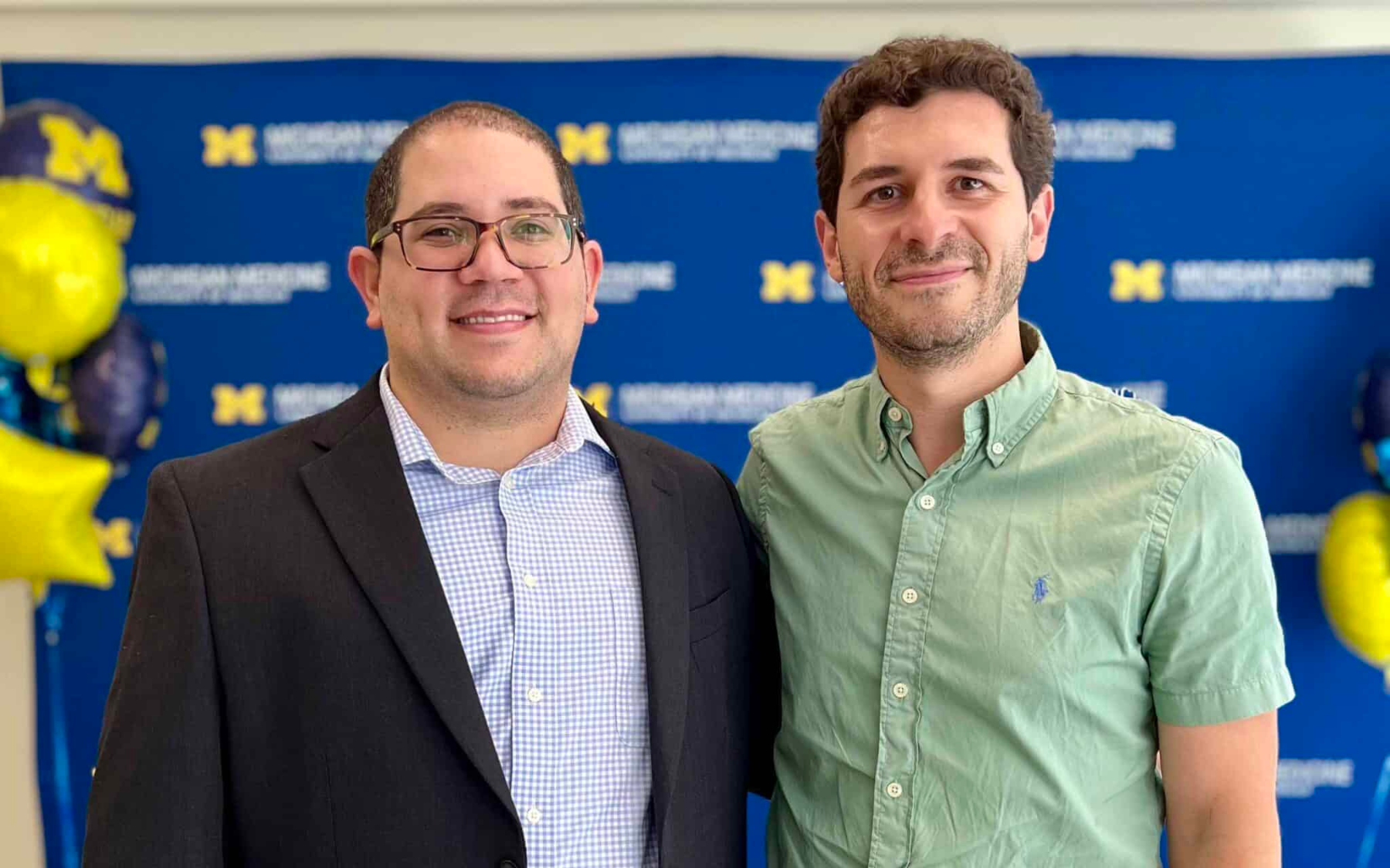 Two men standing in front of a University of Michigan backdrop with balloons. One is wearing a suit, and the other is in a green shirt.