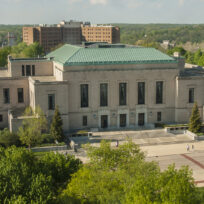 An aerial view of a large classical-style building with a green roof, surrounded by trees, with the University of Michigan Rackham logo in the corner.