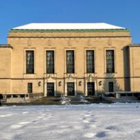 A large beige building with tall windows and a snow-covered foreground under a clear blue sky.