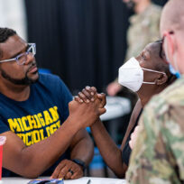 A man and a woman, both wearing masks, hold hands across a table. They are surrounded by military personnel. The man wears a "Michigan Wolverines" shirt.