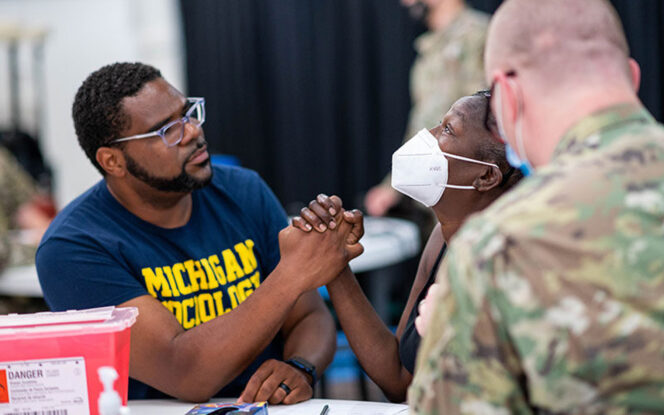 A man and a woman, both wearing masks, hold hands across a table. They are surrounded by military personnel. The man wears a "Michigan Wolverines" shirt.