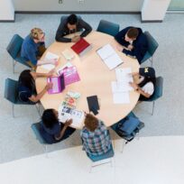 Seven people sitting around a round table with laptops, notebooks, and papers, engaged in a group discussion or study session.