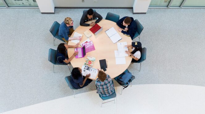 Seven people sitting around a round table with laptops, notebooks, and papers, engaged in a group discussion or study session.