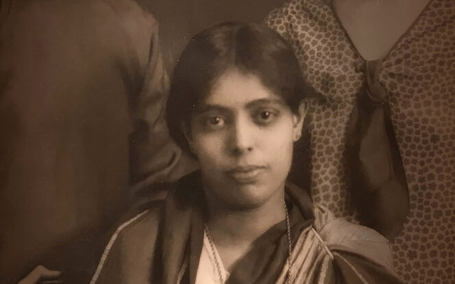 A sepia-toned portrait of a young woman with short hair, wearing a traditional outfit. She is gazing directly at the camera with a neutral expression.