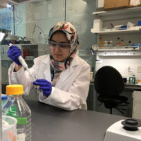 A scientist in a lab coat and gloves uses a pipette at a laboratory bench.
