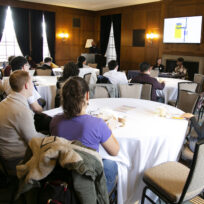 People seated around tables in a conference room, listening to speakers at the front. A screen displays "MSI Summit" above the speakers.