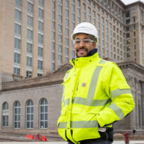 Person wearing a bright yellow safety jacket and hard hat stands in front of a large, historic building.
