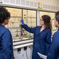 Three people in lab coats stand in a laboratory, discussing chemical equations written on a fume hood.