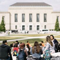 Students gather on a university campus lawn in front of a large, light-colored building with trees and pathways visible.