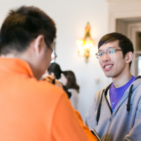 Two people having a conversation indoors. One wears a gray hoodie and glasses; the other is in an orange jacket, facing away. Overhead lights and a doorway are visible.
