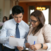 Two people in business attire review documents together in a conference room, surrounded by others engaged in similar activities.