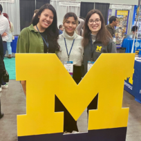 Three people stand behind a large yellow "M" at a university event, with informational booths in the background.