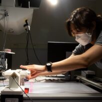 A person wearing a mask adjusts equipment on a lab table under bright lighting.