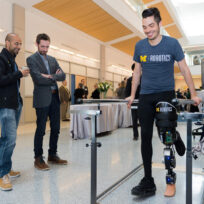 A person with a robotic leg walks using parallel bars while three people observe in a modern indoor setting.