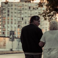 An older couple walks along a street with a tall apartment building in the background, silhouetted by an early evening sky.