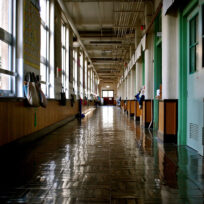 A long, empty school hallway with shiny floors, lined with classrooms and windows on one side, and light streaming in from the windows.