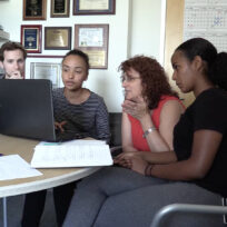 Four people sit around a table with laptops and papers, engaged in discussion. One woman gestures towards the screen while others listen attentively. Plaques and a calendar are visible on the wall.