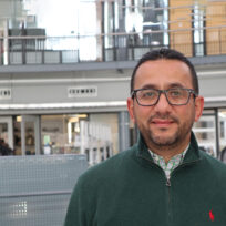 A person with glasses and a beard stands indoors, wearing a green sweater. The background shows a modern, spacious interior with people and architectural details.