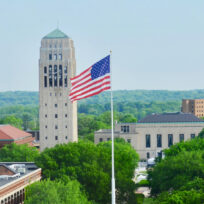 Aerial view of a tall clock tower beside greenery, with an American flag in the foreground and surrounding buildings under a clear sky.