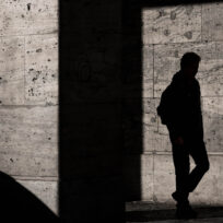 Silhouette of a person with a backpack, standing against a textured wall, partially lit by sunlight. Shadows and geometric shapes from the architecture create contrast.