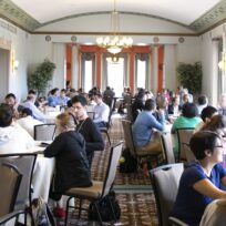 People sitting at round tables in a large, well-lit room, engaged in discussions. The room has chandeliers, large windows, and ornate decor.