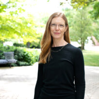 A person with long hair and glasses stands outdoors on a paved path, with green trees and benches in the background.