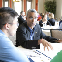 A group of people engage in a discussion around a table in a conference room. One man gestures while speaking. Papers and a pen are visible on the table.