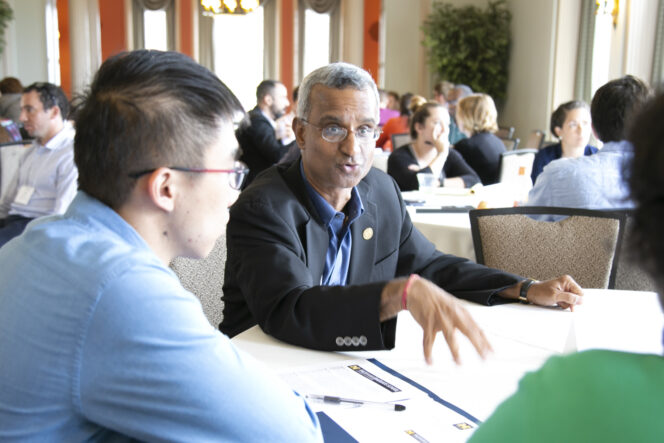A group of people engage in a discussion around a table in a conference room. One man gestures while speaking. Papers and a pen are visible on the table.