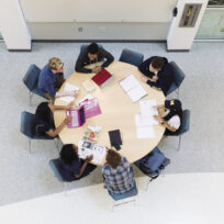 Aerial view of six people sitting around a circular table, engaged in a discussion with laptops and papers.