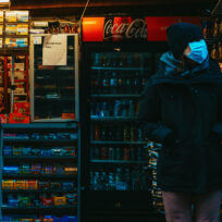 A person wearing a mask stands in front of a well-lit convenience store displaying magazines, drinks, and snacks.