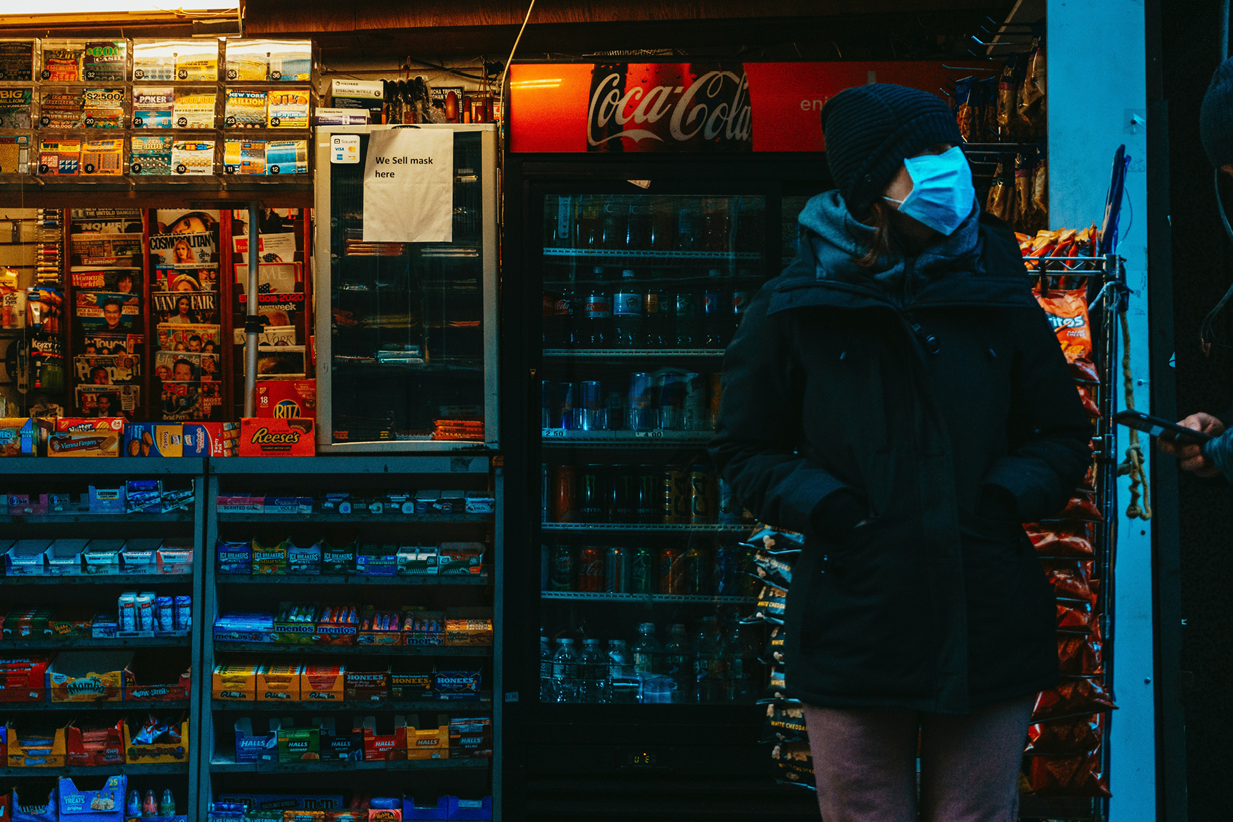 A person wearing a mask stands in front of a well-lit convenience store displaying magazines, drinks, and snacks.