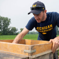 Person wearing a "Michigan Archaeology" shirt sifting soil in a wooden frame in a grassy field.