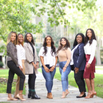 Seven women are posing together outdoors on a pathway, with trees and grass visible in the background.